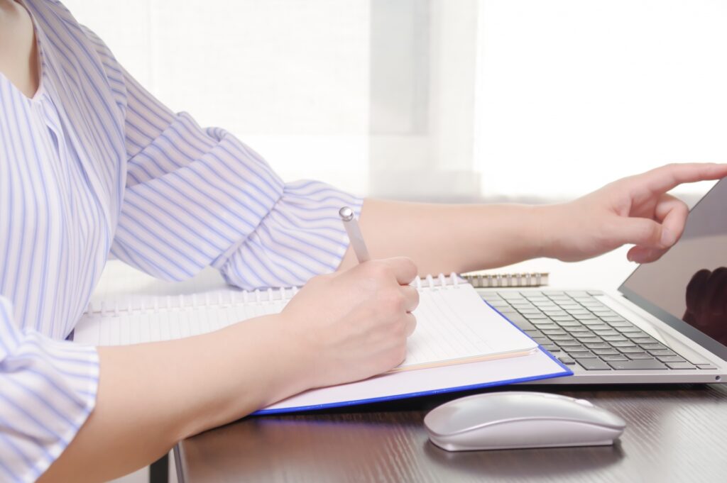 紫のストライプのシャツを着た女性がパソコンを指さしノートをとっている