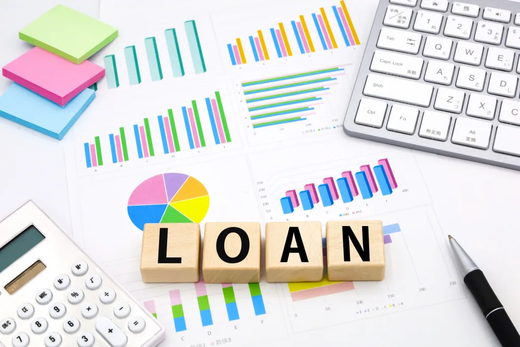 木のブロックにLOANの文字、様々なグラフの書類、付箋、電卓とペン