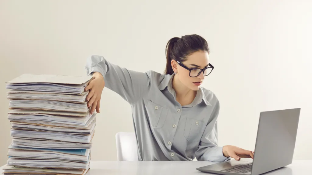 眼鏡をかけた経理の女性が積み上げた書類を横に押しのけてパソコンで作業する