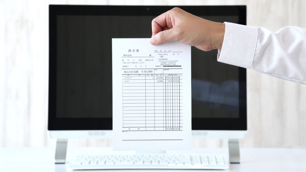 パソコンの前で紙の請求書を持つビジネスパーソン　ペーパーレス化のイメージ